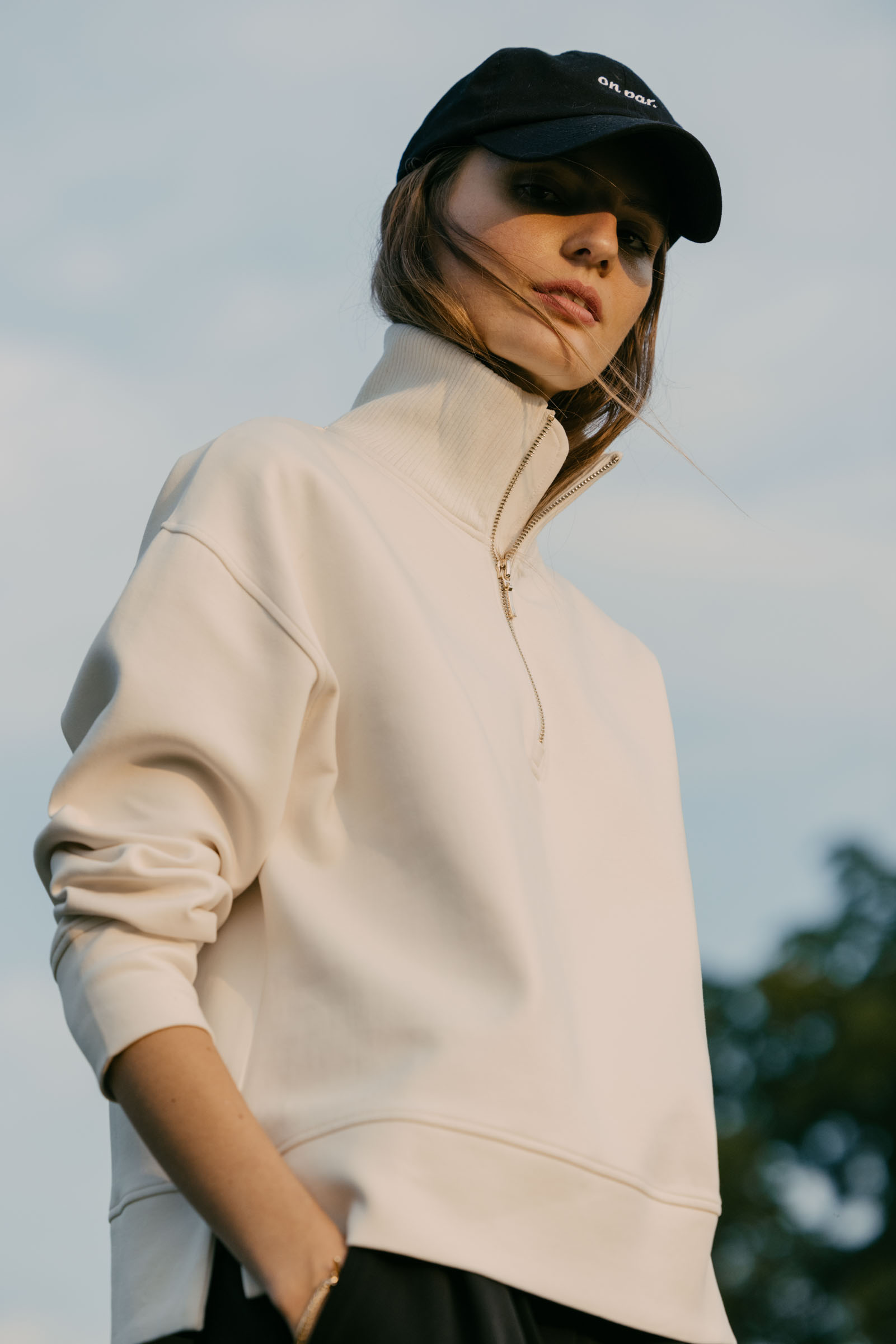 Close up shot of a woman with her hands in her pocket wearing a white quarter-zip and a black baseball cap.