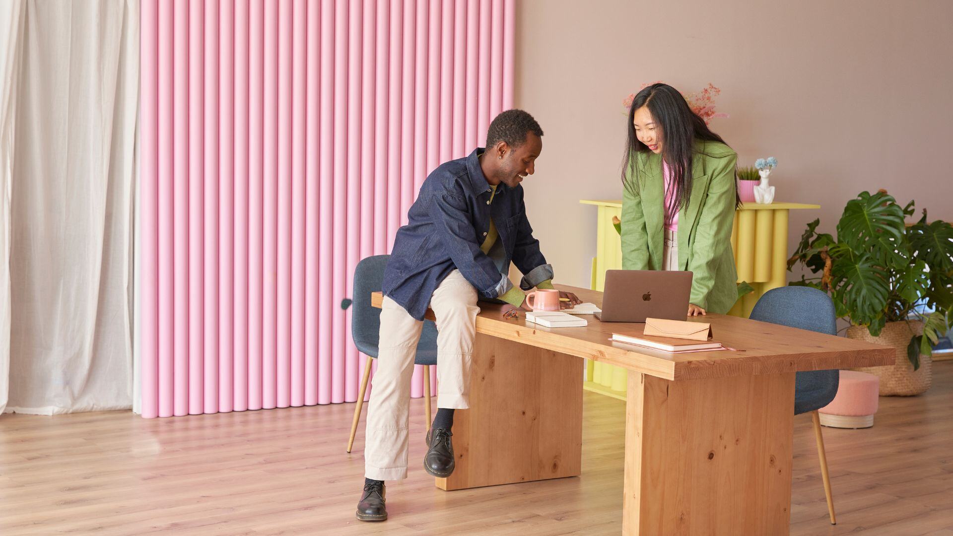 man and women looking at a laptop in a room with pink background and wooden desk