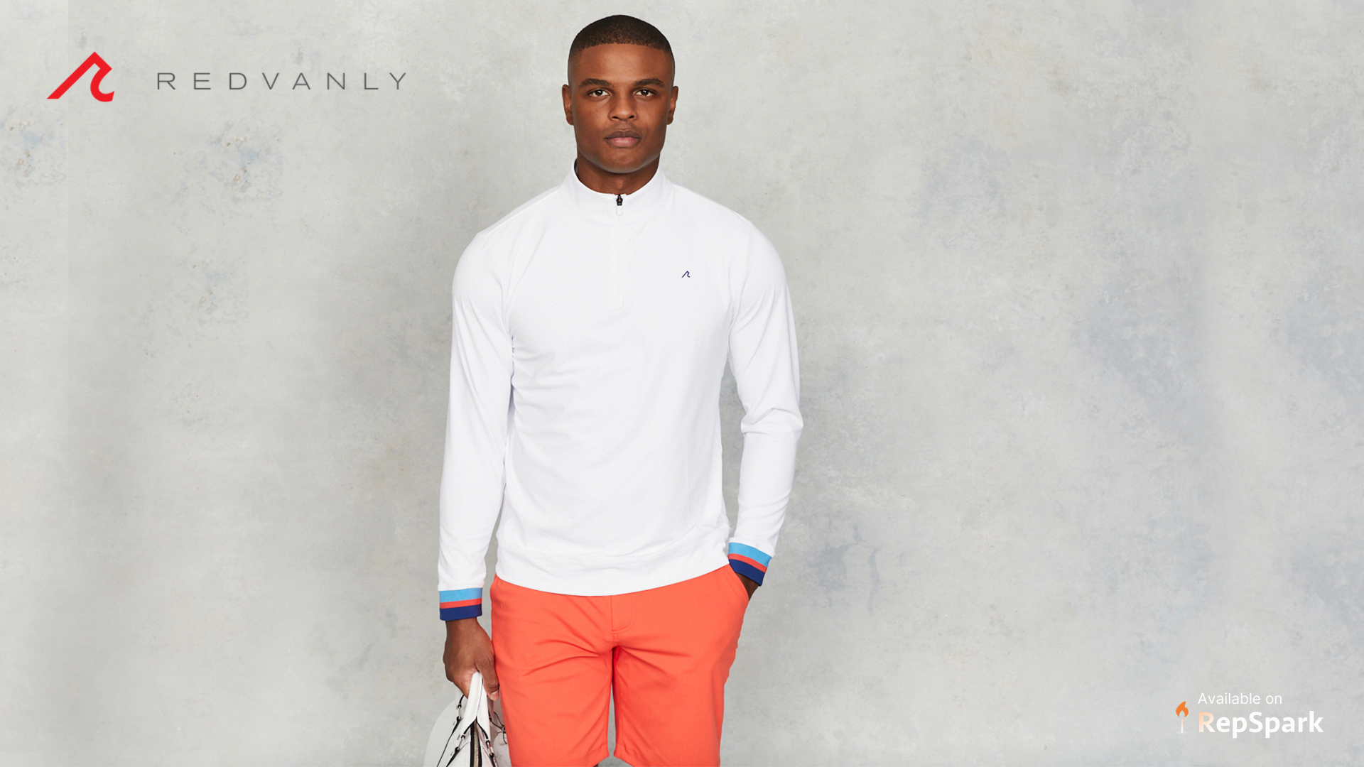 Man in a white long sleeve and coral pants models the brands Redvanly with a grey background.