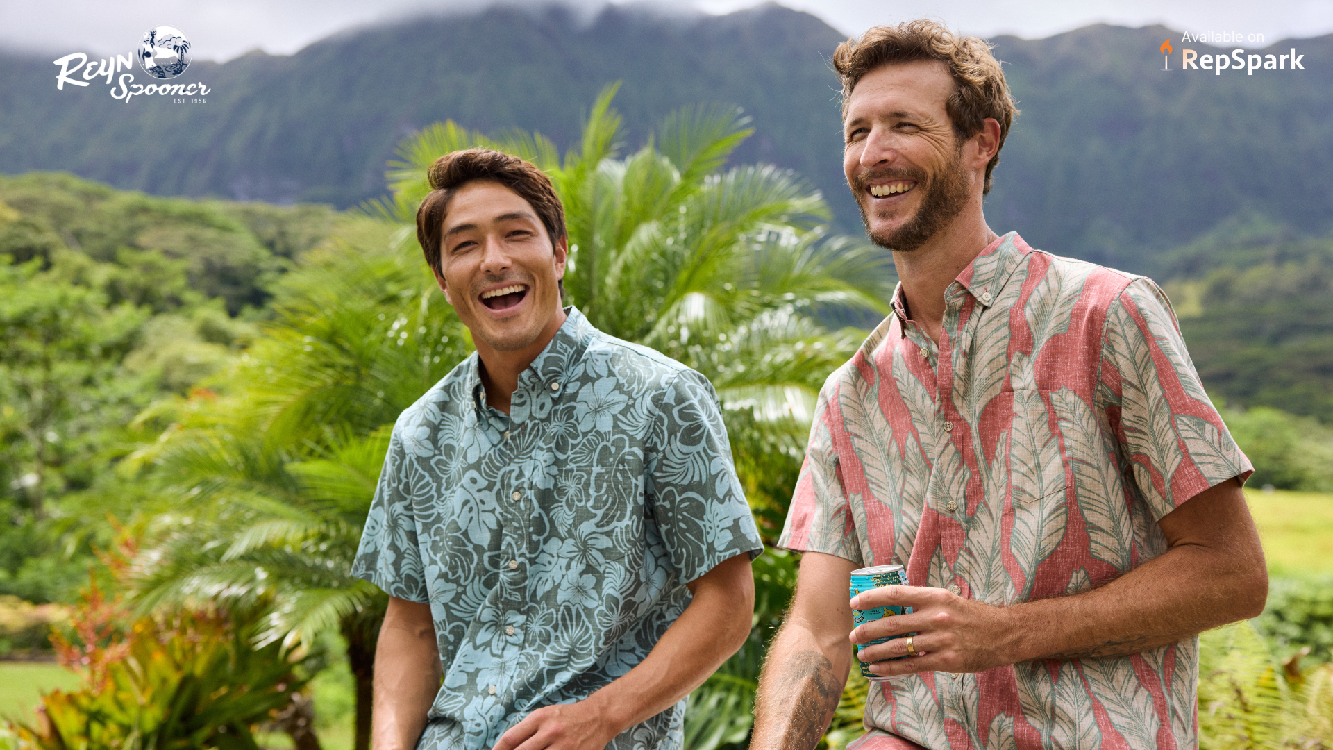 Two men posing together with palm trees in the back. Both men are wearing bright colored Hawaiian t-shits.