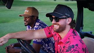 Two men wearing vibrant golf polos and neutral color baseball caps are driving a golf cart on a golf green.